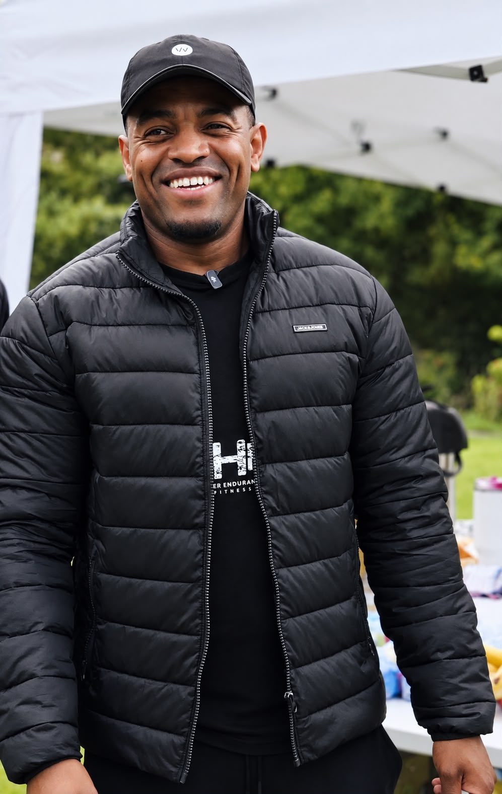 A smiling volunteer in a black cap and puffer jacket over a Hyper Endurance Fitness t shirt beside the food gazebo