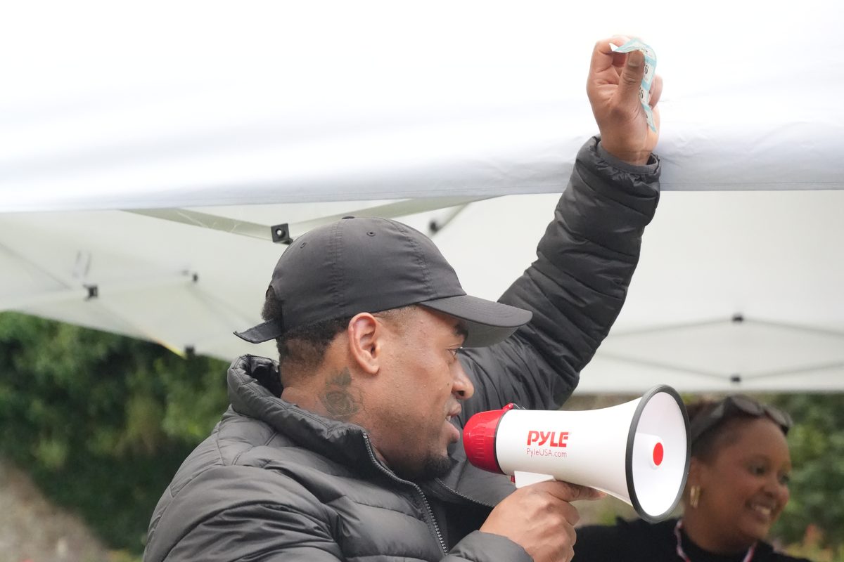 A volunteer in a black cap calling out the raffle ticket numbers through a megaphone under the Community Day gazebo, holding a ticket up in the air